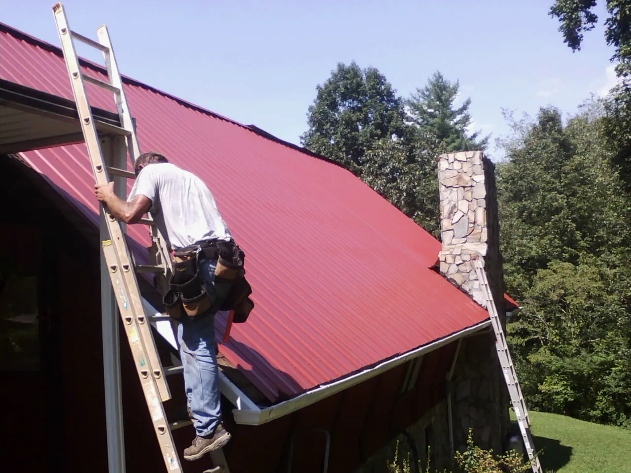 Roofer on a ladder beside a red metal roof and stone chimney — Soffit & Fascia Repair by Roofing Brothers in Chula